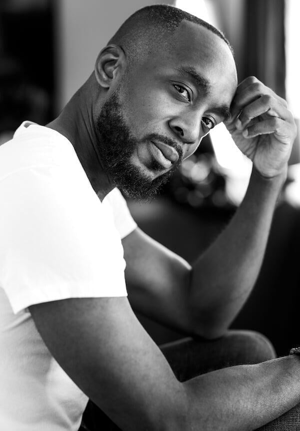 Bearded man, body sideways, head looking to camera.. White shirt. Manicured beard and moustache. Hair cut in tight fade