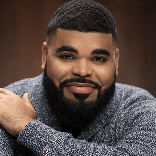 Headshot of Chris Francisque, a Black man with black short hair and beard, smiling at the camera and wearing a great sweater, with his left hand resting on his right shoulder