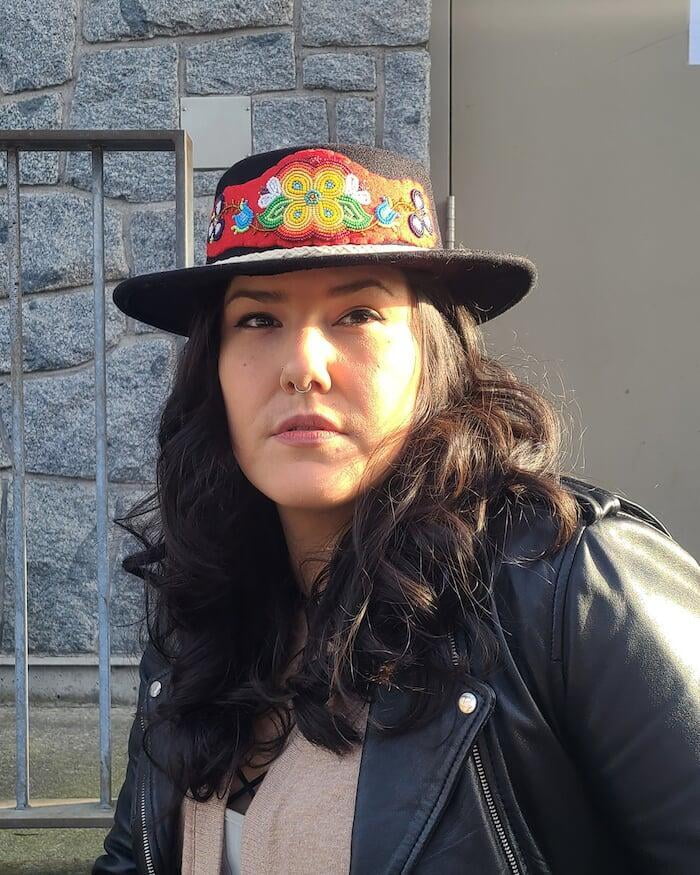 A photo of Debbie with medium length brown hair in a beaded wool hat and a leather jacket in front of a stone wall.