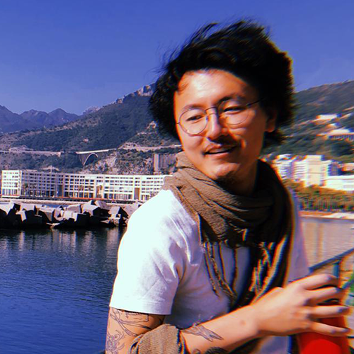 Derek Chan photo, wearing round glasses, a brown scarf and white short-sleeved t-shirt, holding a red cup. In the background are water, mountains, and buildings.  
