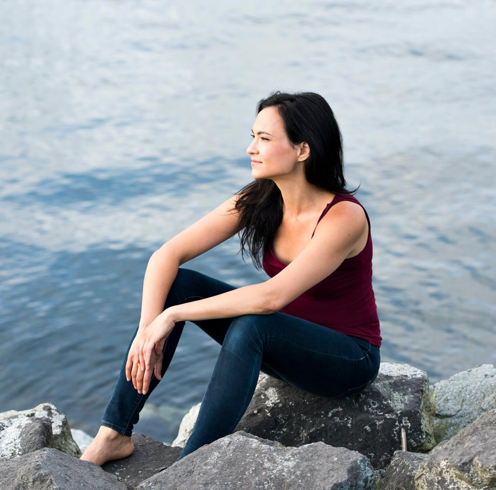 Photo of Emilie Leclerc sitting on a rock at the water's edge. She has long dark hair, is wearing a red tank top and jeans, and resting her elbows on her knees, looking off camera to the left.