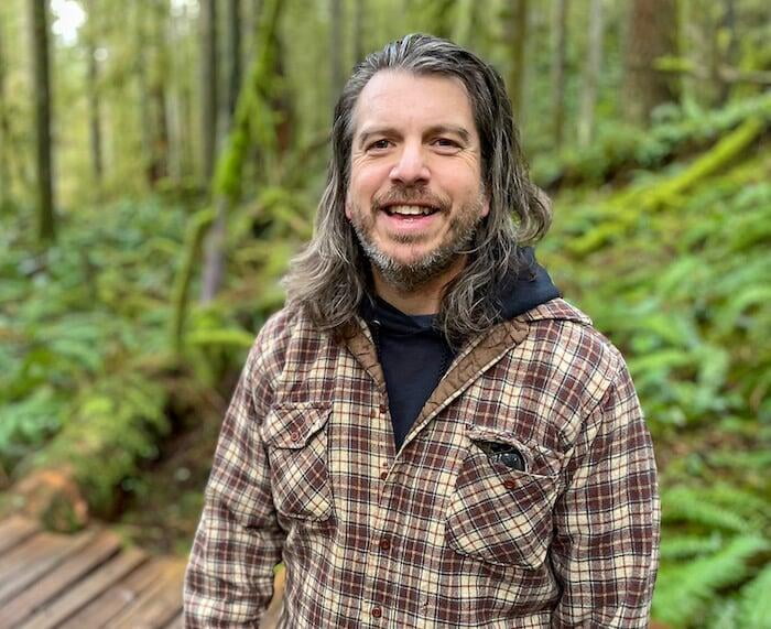 Justin Neal, with brown shoulder length hair and short greying beard, wearing a wool flannel, stands in the woods smiling