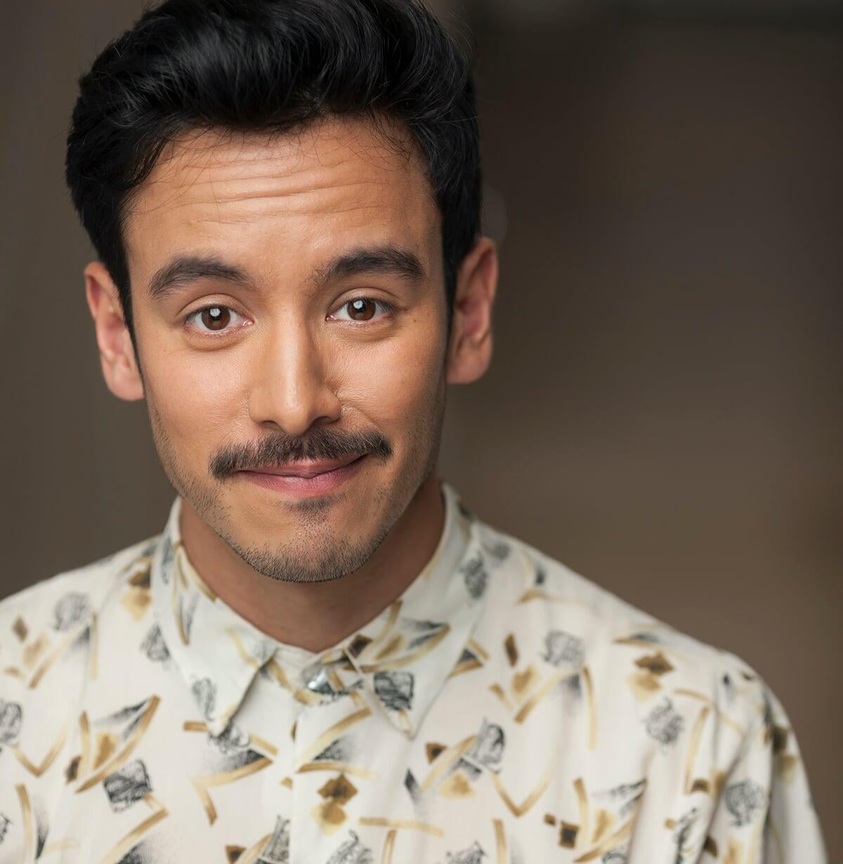 Headshot of Conor Wylie, looking at the camera and slightly smiling, has short dark hair and moustache, dark eyes, wearing patterned collared shirt